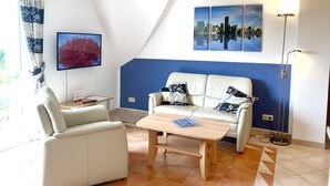 Una sala de estar moderna con un sofá, una mesa de centro y un televisor. Hay tres cuadros en la pared y una lámpara de pie.