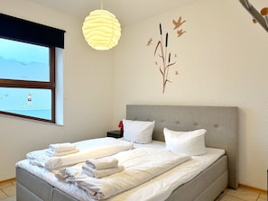 Una cama doble con sábanas blancas, cabecera gris y lámpara de pared.