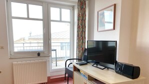 A small room with a desk, a television, and a chair. There is a window with a view of buildings outside.