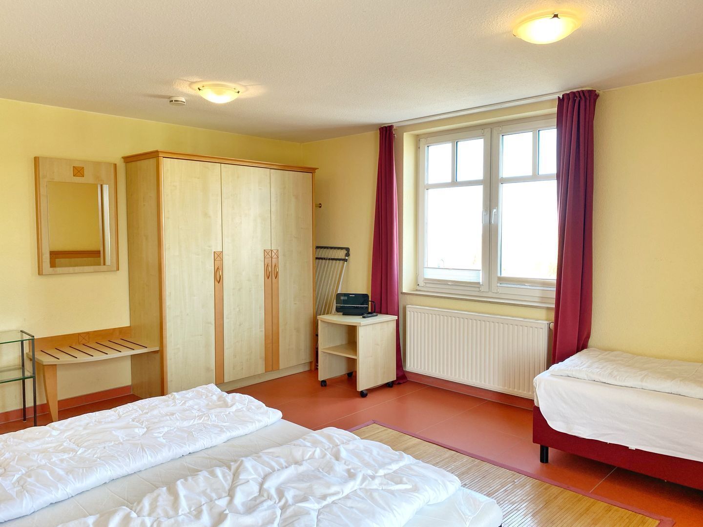 Ein Schlafzimmer mit zwei Betten, einem Schrank, einem Schreibtisch, einem Stuhl, einem Heizkörper und einem Fenster mit Vorhängen.