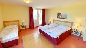 Une chambre d’hôtel avec deux lits simples, un grand lit, une fenêtre avec des rideaux, un tableau au mur et une petite table avec une lampe.