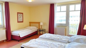 Une chambre d’hôtel avec deux lits simples, un lit double, une table de chevet avec une lampe, un radiateur et une fenêtre avec des rideaux.