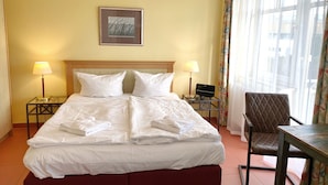 A hotel room with a bed, two bedside lamps, a small table, a chair, and a framed picture on the wall.