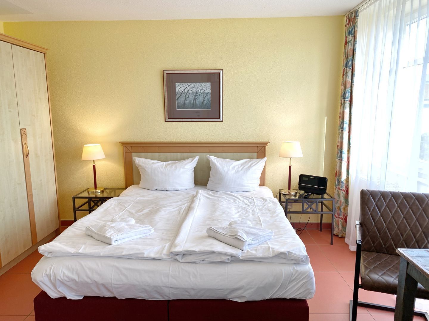 Habitación de hotel con cama, dos lámparas de noche, un cuadro en la pared y una silla junto a la ventana.