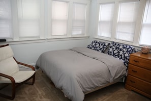 Une chambre à coucher avec un lit, une chaise, une commode et des fenêtres équipées de stores.