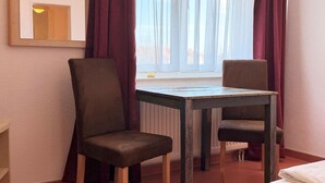 Ein Zimmer mit Holztisch, zwei Stühlen, einem Bett, einem Fenster mit Vorhängen und einem Spiegel an der Wand.
