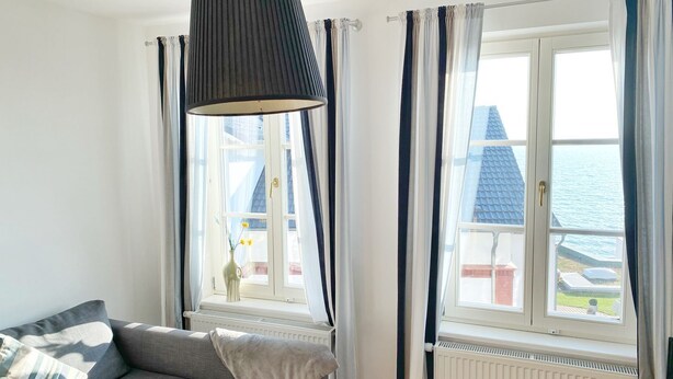 Ein Zimmer mit grauem Sofa, einer Pendelleuchte und Meerblick durch große Fenster.