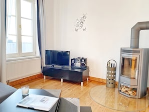 Un salon moderne avec un téléviseur à écran plat, un canapé noir, une table basse en verre avec un magazine, un élément décoratif mural et un poêle à bois.