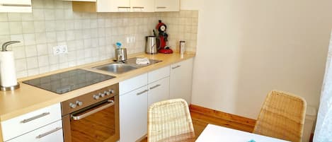 A compact kitchen with white cabinets, a stainless steel stove, and a sink. There are wicker chairs and a table with a blue placemat.