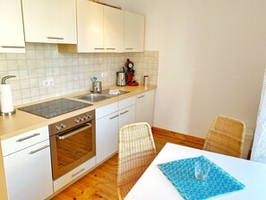 A compact kitchen with white cabinets, a stainless steel stove, and a sink. There are wicker chairs and a table with a blue placemat.