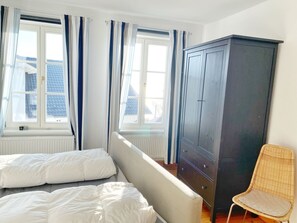A bedroom with two beds, a wardrobe, a chair, and large windows with curtains. - Seaside Appartements, "Grey" - Sassnitz - Seaside Appartements (Sassnitz)
