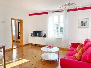 A bright living room with a wooden floor, a white cabinet with a TV, a red sofa, and a small white table. - Seaside Appartements, "Red" - Sassnitz - Seaside Appartements (Sassnitz)