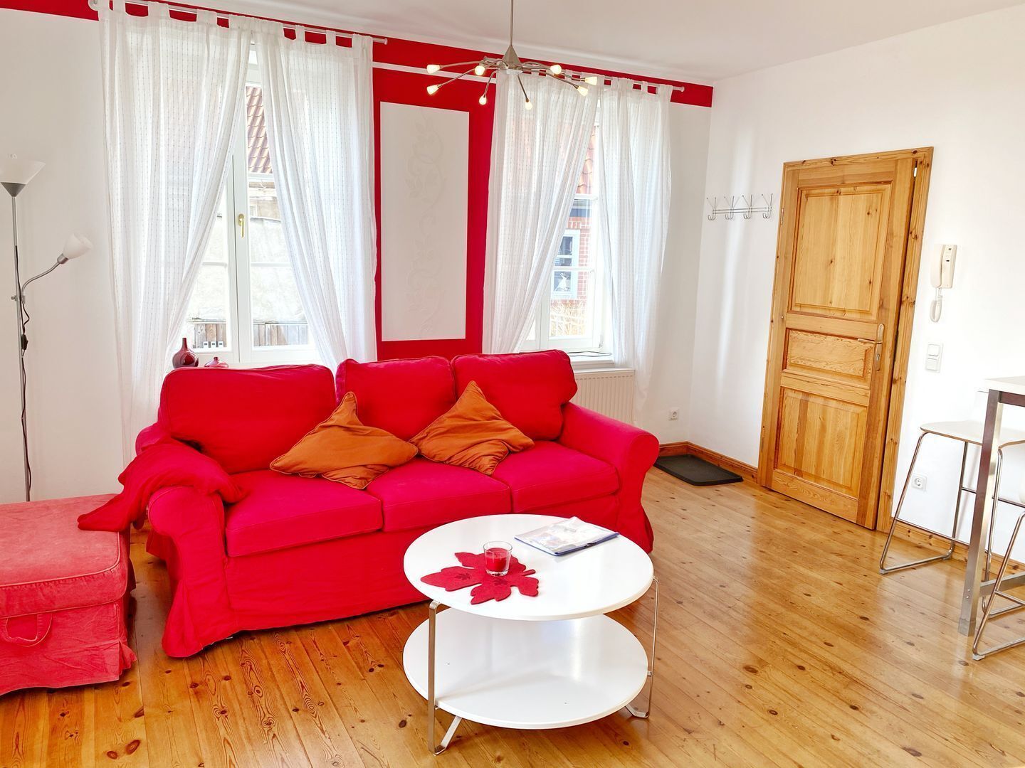 Ein rotes Sofa mit zwei orangefarbenen Kissen, ein weißer runder Tisch mit einer roten Blumendekoration und eine Holztür.