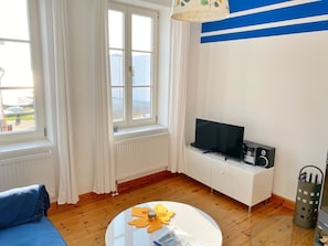 A room with a TV, a white cabinet, a blue sofa, a round table with a candle, and a window with curtains.
