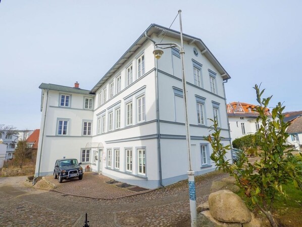 A multi-story building with a white facade and blue trim, a cobblestone driveway, and a parked car.