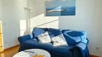 Ein blaues Sofa mit gemusterten Kissen, ein weißer Couchtisch mit einem Glas und orangefarbenen, blattformigen Büroklammern, und ein Gemälde einer Küstenlandschaft an der Wand.