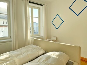 A bedroom with two single beds, a radiator, a window with curtains, and a chair.
