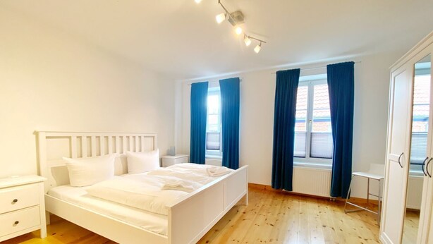 Une chambre avec un lit blanc, un plancher en bois et une fenêtre ornée de rideaux bleus.