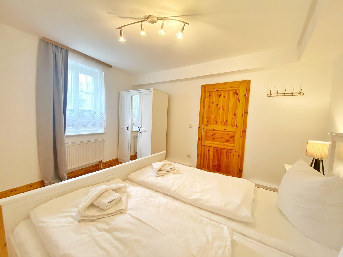 A bedroom with two beds, a wooden door, a window with curtains, a radiator, and a ceiling light fixture.