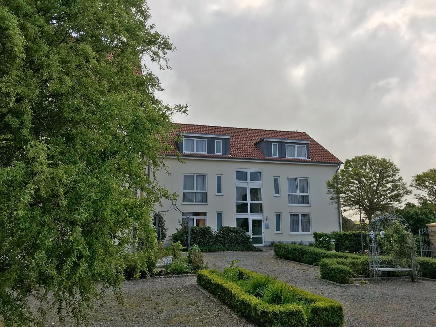 Un bâtiment à deux étages, doté d’un toit recouvert de tuiles rouges, de nombreuses fenêtres et d’un jardin bien entretenu avec des haies et un chemin de gravier.
