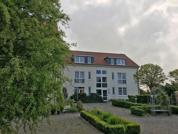 A two-story building with a red-tiled roof, multiple windows, and a well-maintained garden with hedges and a gravel path.