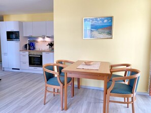 A compact kitchen with white cabinets, a microwave, and a coffee maker. There is a dining table with four chairs and a painting of a beach scene on the wall.