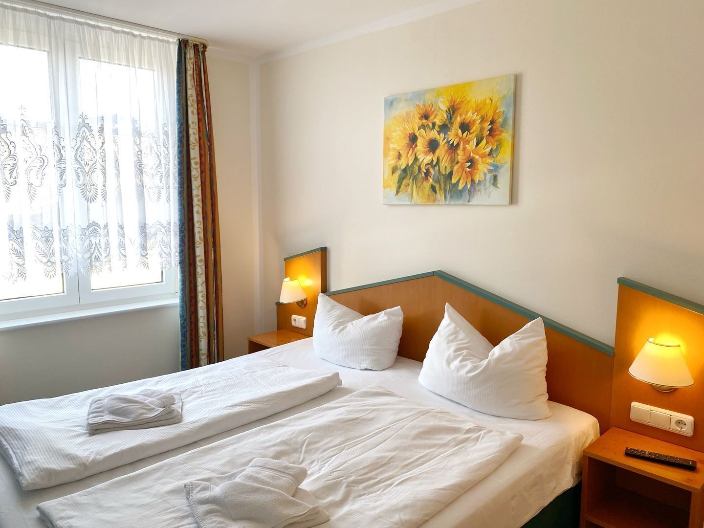 Ein Hotelzimmer mit einem aufgeräumten Bett, zwei Nachttischlampen, einem Sonnenblumenbild an der Wand und einem Fenster mit Vorhängen.
