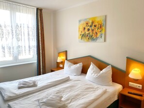 Ein Hotelzimmer mit einem aufgeräumten Bett, zwei Nachttischlampen, einem Sonnenblumenbild an der Wand und einem Fenster mit Vorhängen.