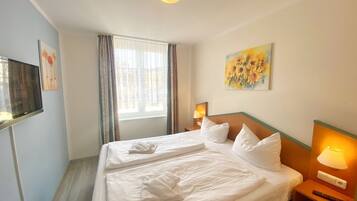 Et hotelværelse med en stor seng, et fjernsyn til venstre, et vindue med gardiner, et billede på væggen og en sengetavlelampe.