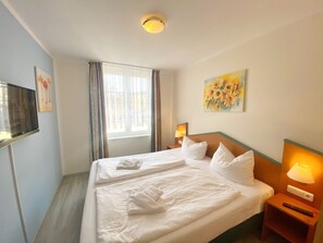 A hotel room with a large bed, a TV on the left, a window with curtains, a painting on the wall, and a bedside lamp.