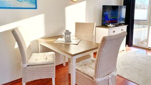 Ett rum med ett matbord i trä, stolar av rotting, en tv-apparat på ett skåp och ett stort fönster med gardiner.