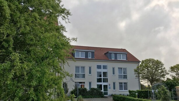 Een gebouw met twee verdiepingen, voorzien van een rood pannendak, meerdere ramen en een verzorgde tuin met heggen en een grindpad.