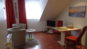 Een kleine, moderne kamer met een schuine kap, een flatscreen-tv, een bureau, een stoel, een bank en een raam met rode gordijnen.