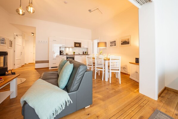 A modern living room with a grey sofa, a dining area with white chairs, and a kitchen with white cabinets.