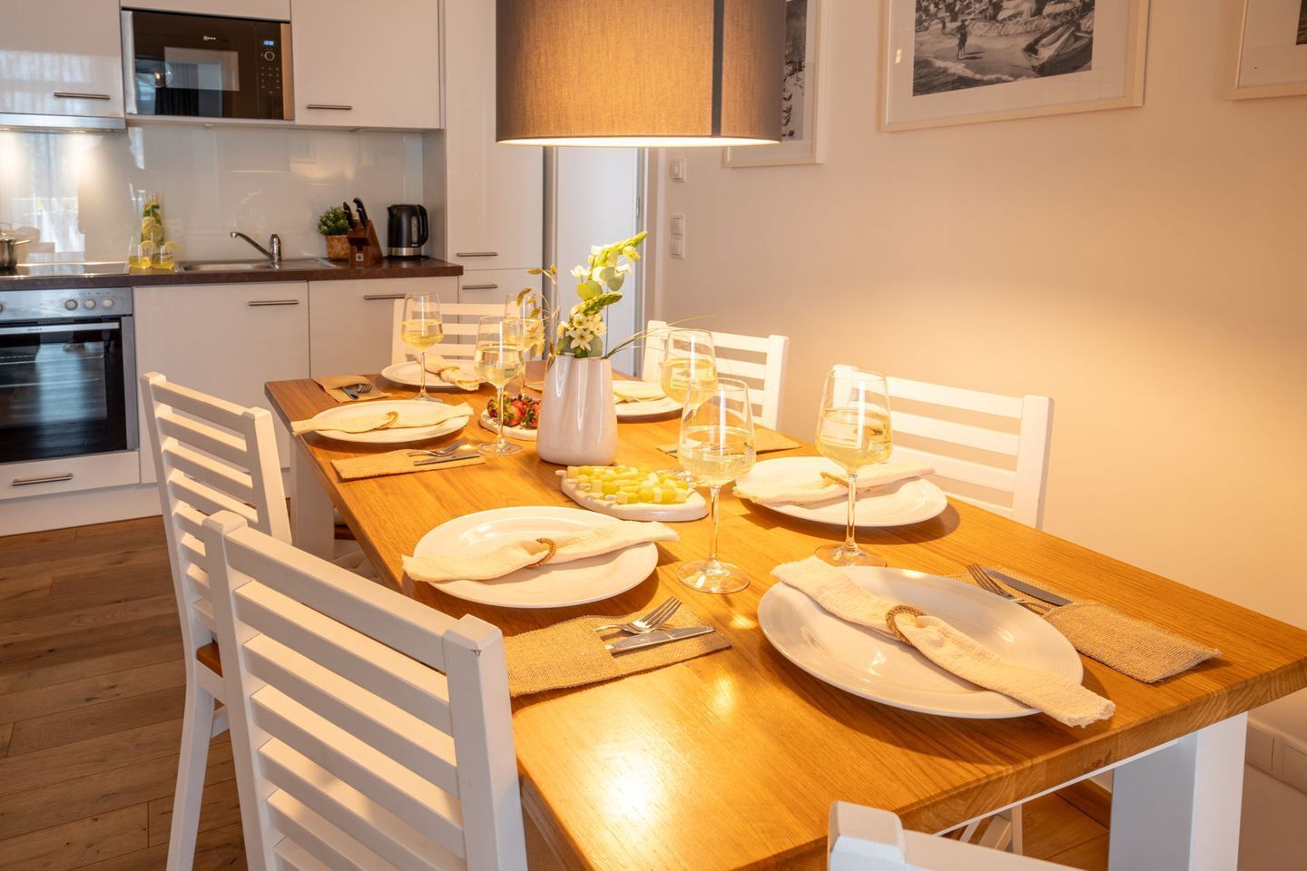 A modern kitchen with white cabinetry, a wooden dining table set for four, and a large window above the table.
