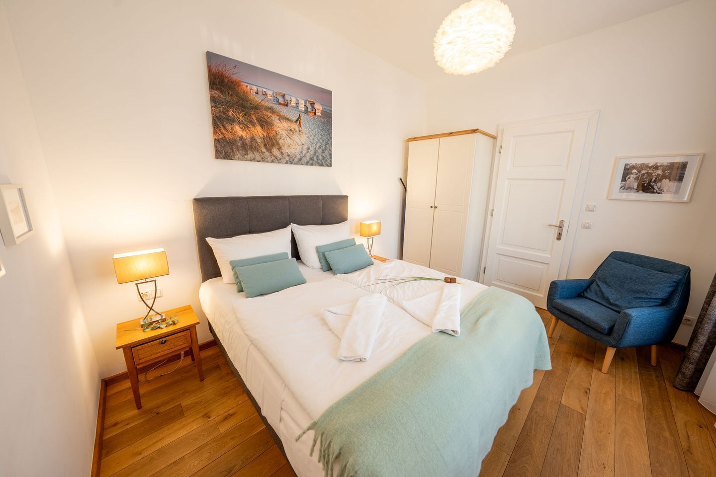 A bedroom with a bed, bedside lamps, a chair, a wardrobe, and a painting on the wall.