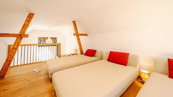 A high-ceilinged room with wooden beams, two beds with red pillows, a wooden floor, and a balcony with a railing.