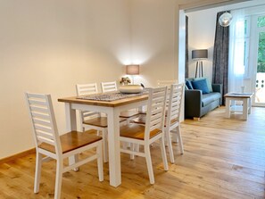 Uma área de refeições com mesa de madeira, cadeiras brancas, uma mesinha lateral e um sofá ao fundo.