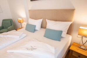 A hotel room with a bed, two pillows, a nightstand, a lamp, a chair, and a towel with a logo.