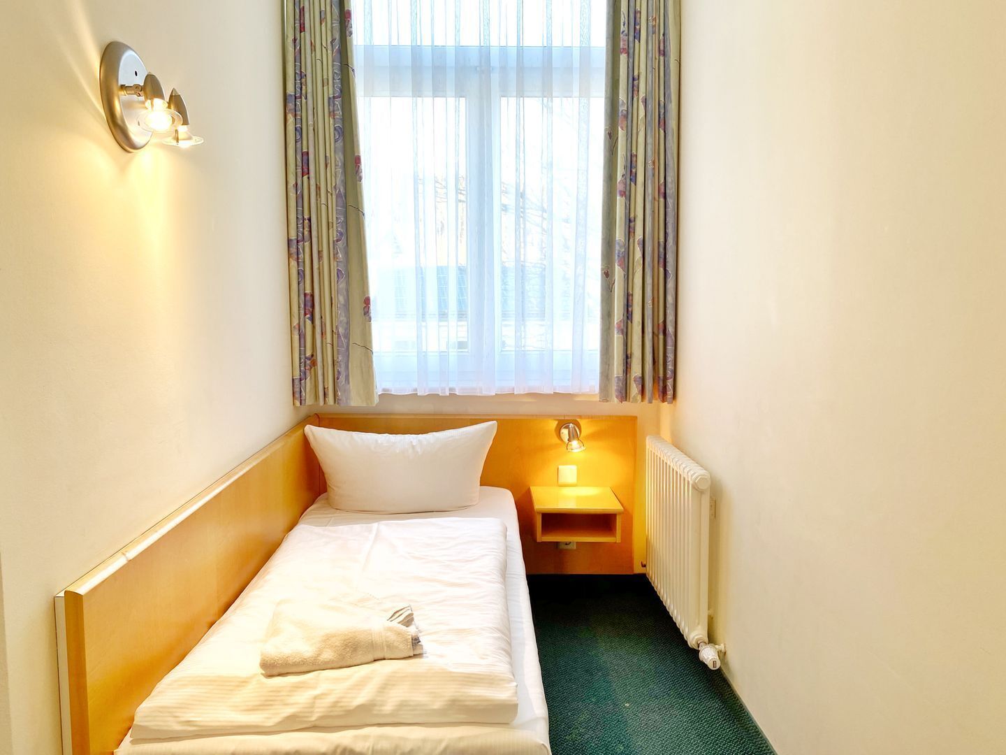 A small, single-bed room with a wooden headboard, a bedside table with a lamp, a radiator, and a window with curtains.