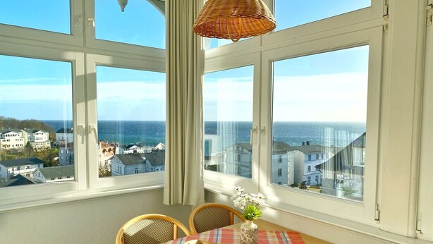 A dining area with a table and chairs, a wicker hanging light, and a view of the sea and buildings. - Sea view apartment no. 21 in the Villa Anna in Sassnitz on Rügen - Sassnitz - (Sassnitz)