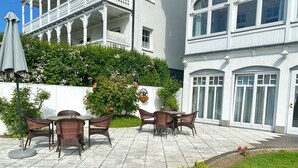 An outdoor patio with wicker furniture, a white building with a balcony, and a garden area with flowers. - Sea view apartment no. 21 in the Villa Anna in Sassnitz on Rügen - Sassnitz - (Sassnitz)