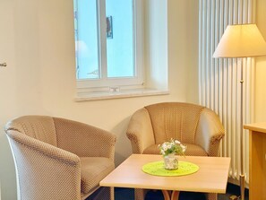 Um quarto com duas poltronas estampadas, uma mesa pequena com um vaso de flores, uma janela com persianas e um abajur.