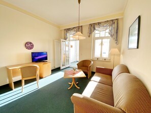 Ein Hotelzimmer mit einer Couch, einem Sessel, einem Schreibtisch mit Fernseher, einer Lampe und einem Fenster mit Vorhängen.