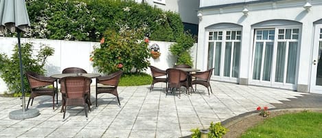 An outdoor patio with wicker furniture, a white building with a balcony, and a garden area with flowers.