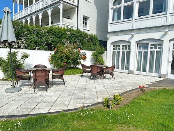 An outdoor patio with wicker furniture, a white building with a balcony, and a garden area with flowers.