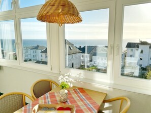A room with a table set for two, a wicker lamp, and a view of the sea and buildings through large windows. - Sea view apartment no. 23 in the Villa Anna in Sassnitz on Rügen - Sassnitz - (Sassnitz)