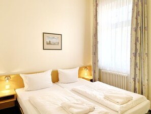 Ein Doppelbett mit weißer Bettwäsche, zwei Nachttische mit Lampen, ein gerahmtes Bild an der Wand und ein Fenster mit floralen Vorhängen.