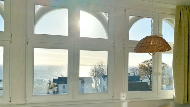 A room with large arched windows, a wicker lamp, a table with a plaid tablecloth, and a view of the sea and buildings. - Sea view apartment no. 32 in the Villa Anna in Sassnitz on Rügen - Sassnitz - (Sassnitz)