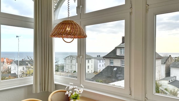 Una habitación con ventanas amplias que ofrecen vista al mar y edificios. Se observa un colgante de luz de mimbre, una mesa con mantel de cuadros y un florero con flores.